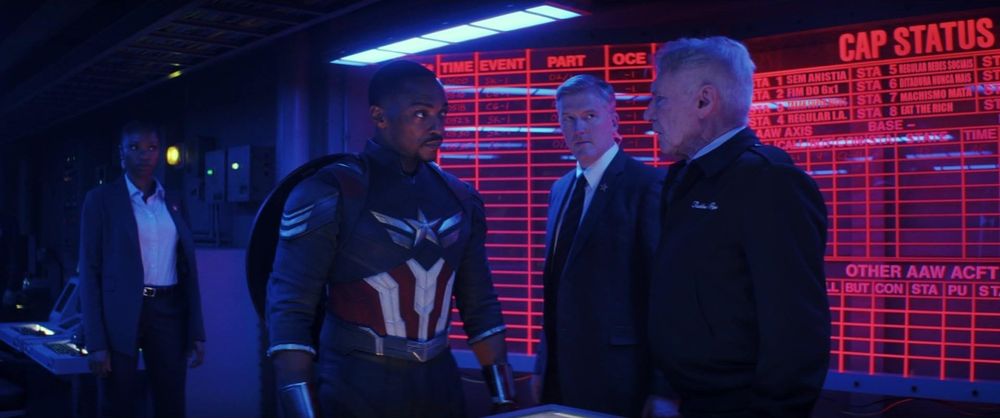 Em uma sala de comando iluminada por luz azul e vermelha, um homem vestindo um uniforme de super-herói azul, vermelho e branco conversa seriamente com dois homens mais velhos que usam roupas formais, um deles com uniforme militar. Ao fundo, há um grande painel eletrônico vermelho com diversas linhas e textos. À esquerda, uma mulher de terno observa a cena.