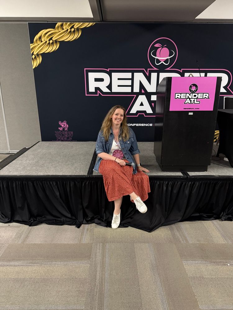 Photo of Kelly sitting on stage at RenderATL holding a microphone 