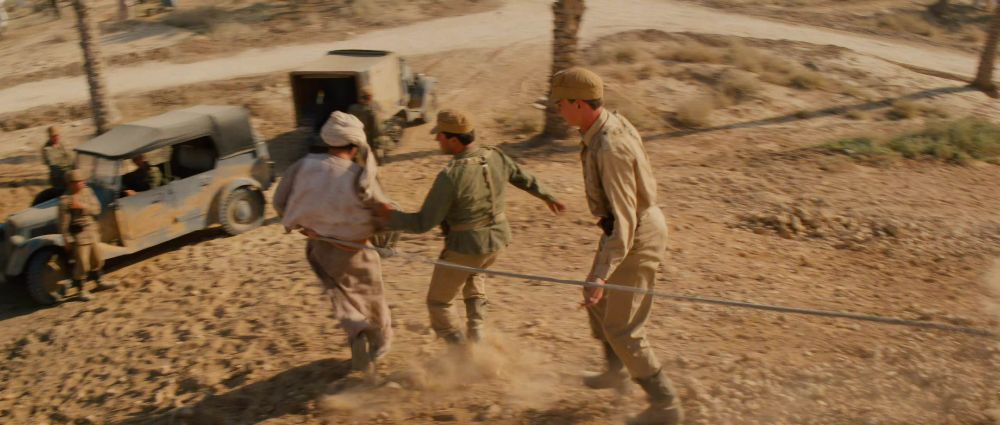 Still from Raiders of the lost Ark: The soldiers lead Sallah down the hill, away from the map room. At the bottom of the hill there are two military vehicles.