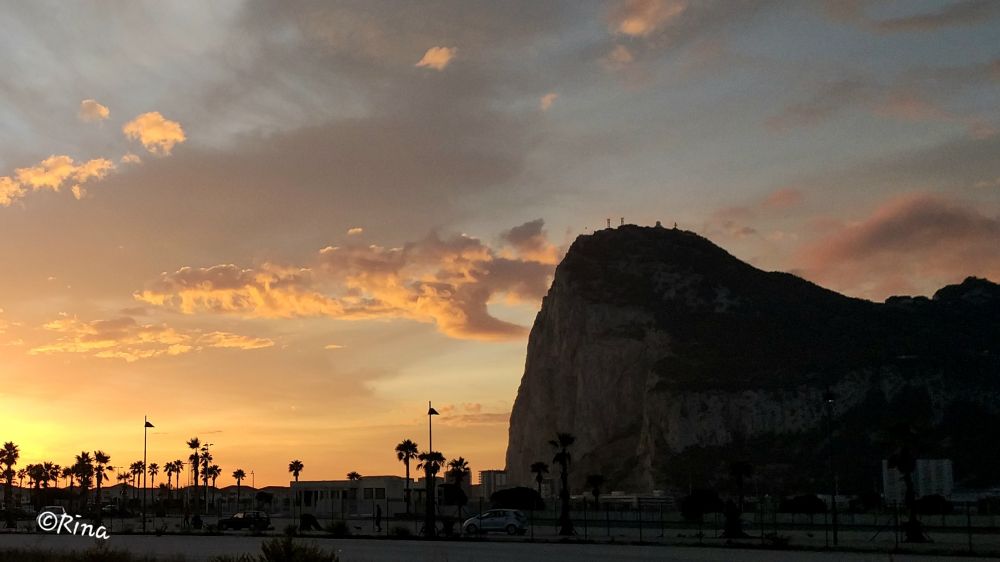 One can see the Rock of Gibraltar at sunset. The rock is on the right side, on the left the sky is yellow from the setting sun, and there are pink glowing clouds in the sky. In the foreground, one can see a road and the silhouettes of palm trees.