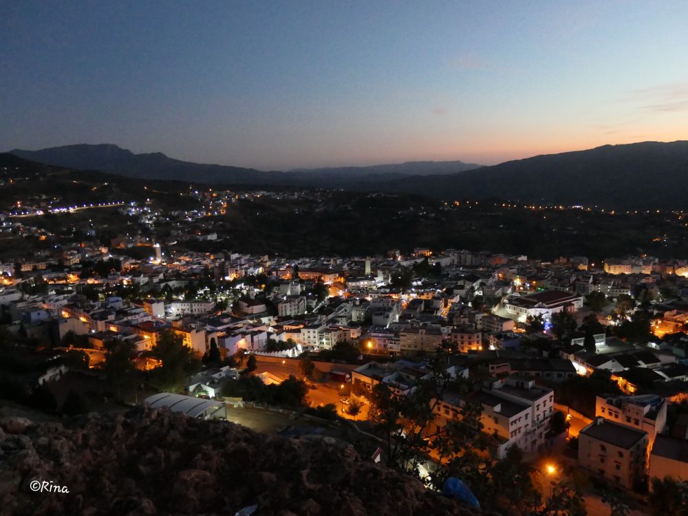 You look from a hill at an illuminated city. The sky over the Rif Mountains is still lit up blue after sunset