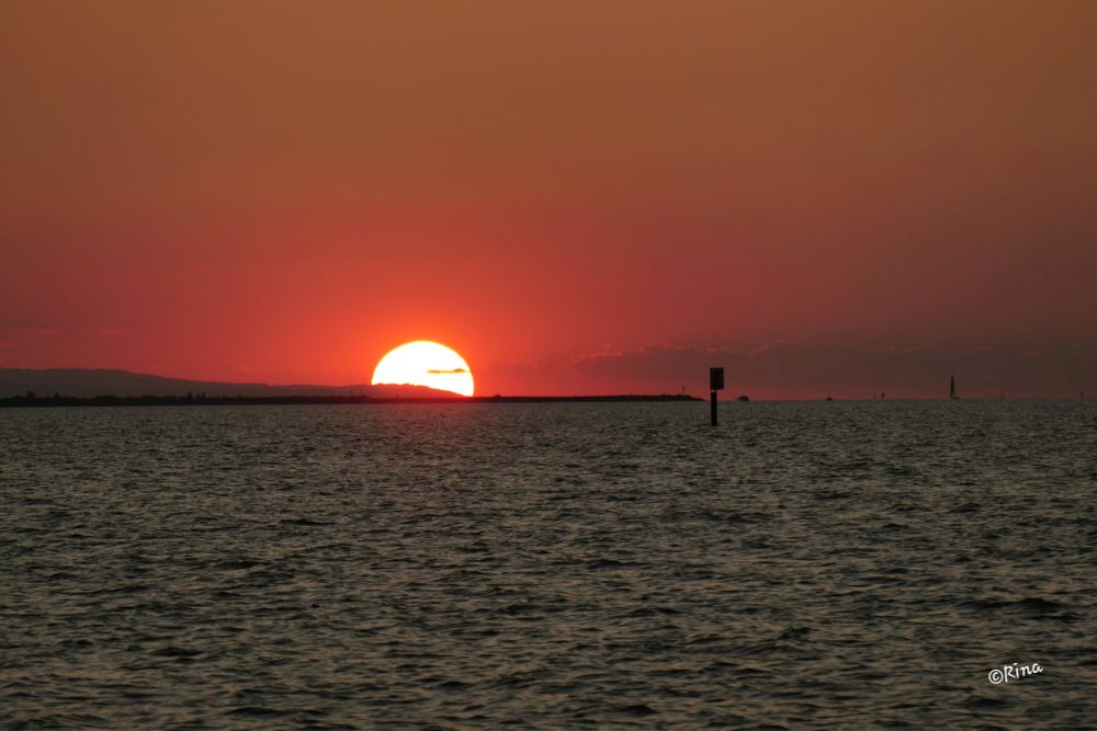 The sun sets as a red fireball behind the Rhine dam. The sky is orange, in the foreground is the water of Lake Constance and seamarks