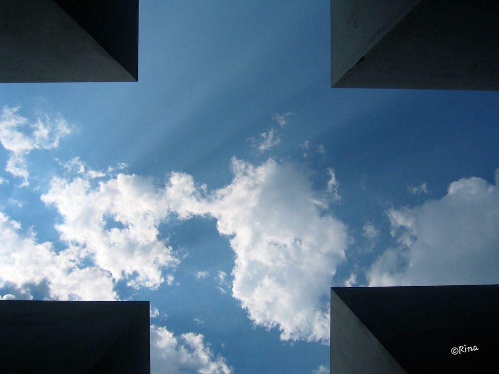 Standing between concrete pillars, a photo of the sky was taken. In each corner of the photo, a dark pillar can be seen, creating a cross in which the blue sky with white clouds is visible. Sun rays can be seen.