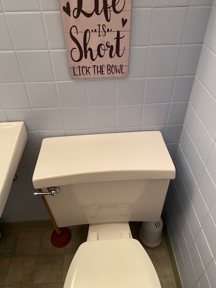 A toilet. Above it, on the wall, is a wooden inspirational poster-style hanging reading “Life is short, lick the bowl”