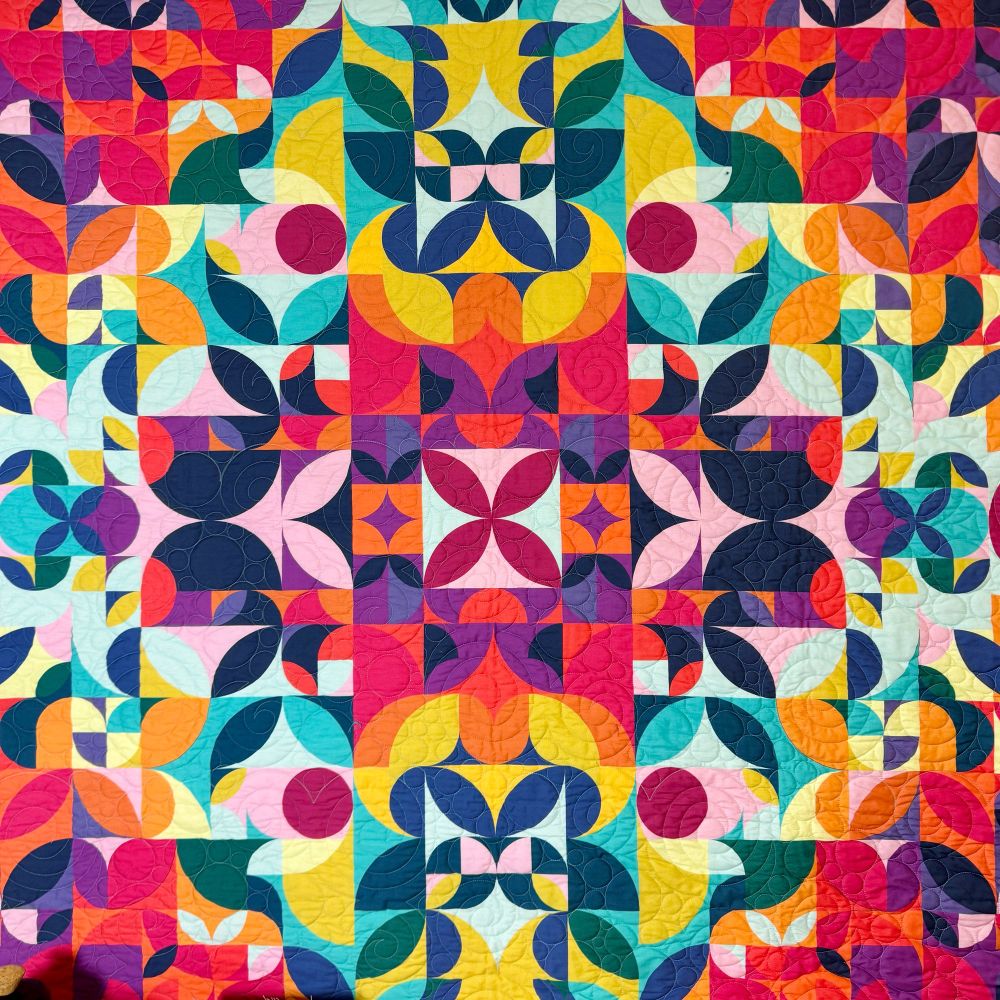 A photo of a colourful kaleidoscopic quilt made up of many curves. Coloured in reds, purples, pinks, blues and yellows.