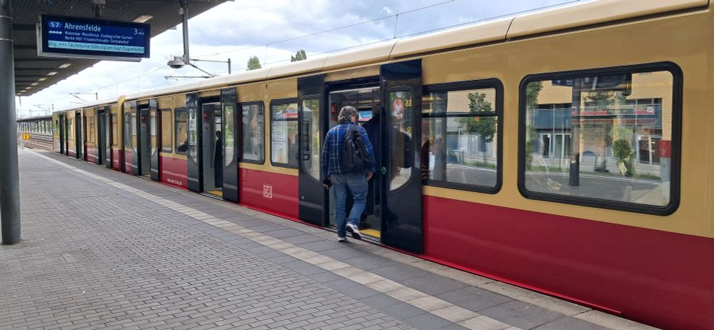 Eine S-Bahn (oben beige, unten rot) steht an einem Bahnsteig. Passagiere steigen ein.