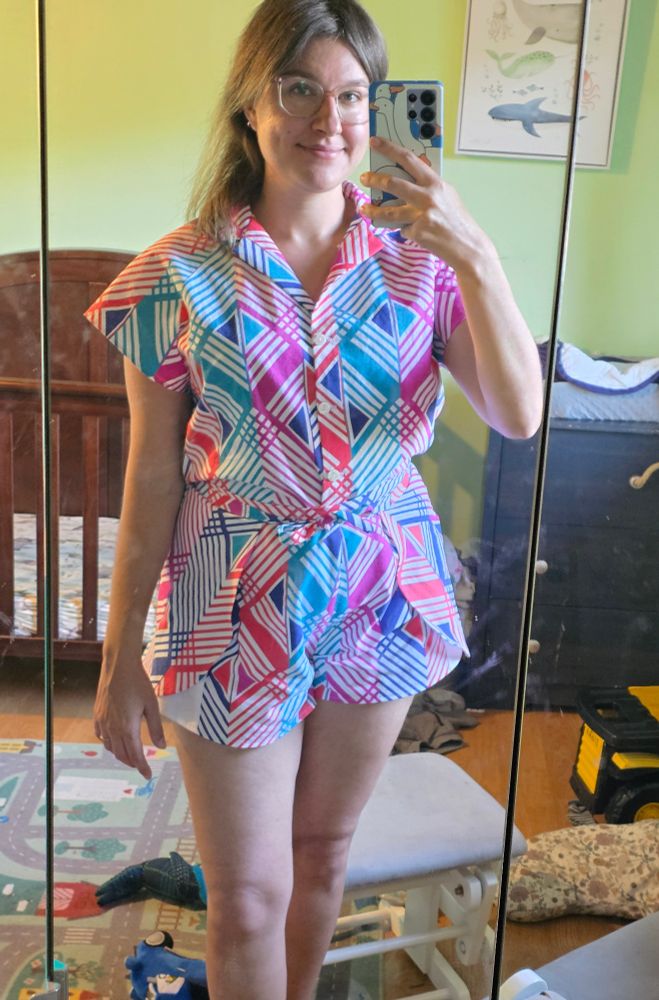 Mirror picture of me in a button up and wrap shorts, baby room in the background. 