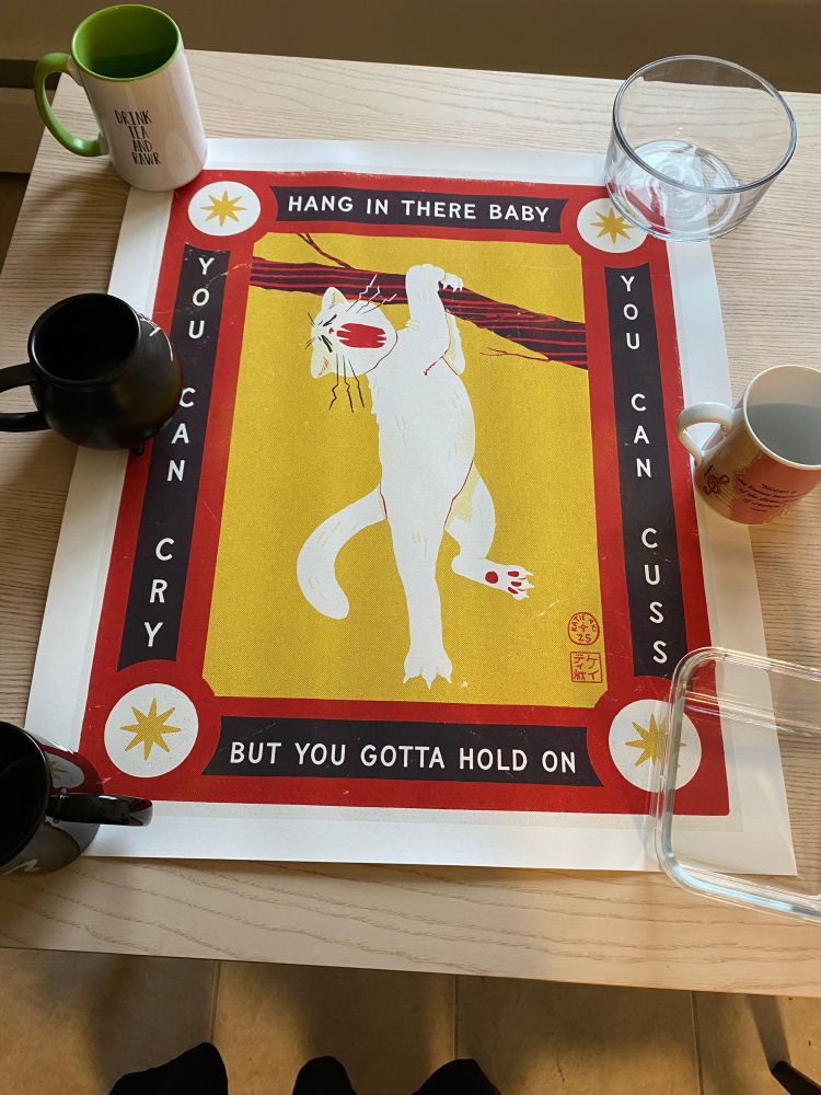 Print by Katie Aiki - Cat hanging from a branch with the words "HANG IN THERE BABY/YOU CAN CRY/YOU CAN CUSS/BUT YOU GOTTA HOLD ON" around edges. Colours are red, white, yellow, black.