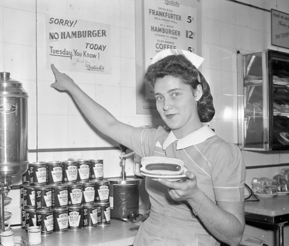 Black and white photo of a waitress holding a hot dog in the 1940s. She's pointing at a sign that says:
SORRY!
NO HAMBURGER TODAY
Tuesday, You Know !