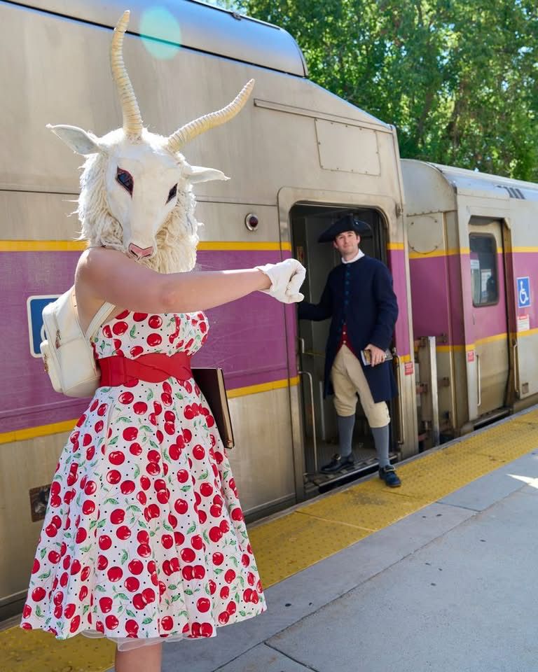 Lady in a horned sheep head and patterned dress pointing at a guy in colonial garb getting on an mbta train