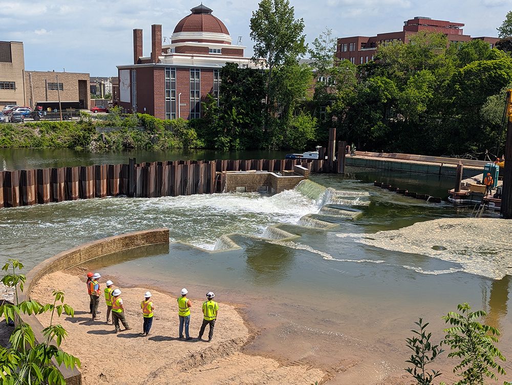 Looking north at water flowing over the labyrinth weir, part of the FishPass facility on the Boardman Ottaway River in Traverse City, Michigan. Workers are watching a crane removing the barriers in the river to restore the flow. They'll be closing off flow on the northern section to begin work on the fish sorting channel.