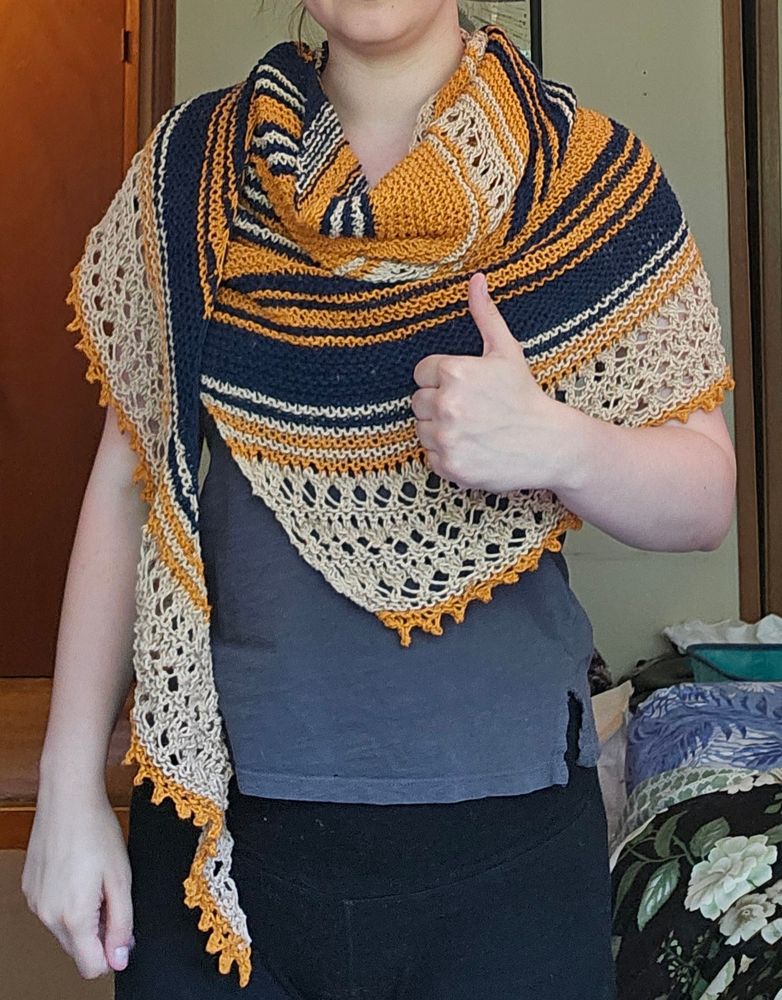 A person giving the camera a thumbs up, wearing a knit shawl in orange, dark blue, and beige in alternating stripes of varying size. There's some lace detailing on the edges.