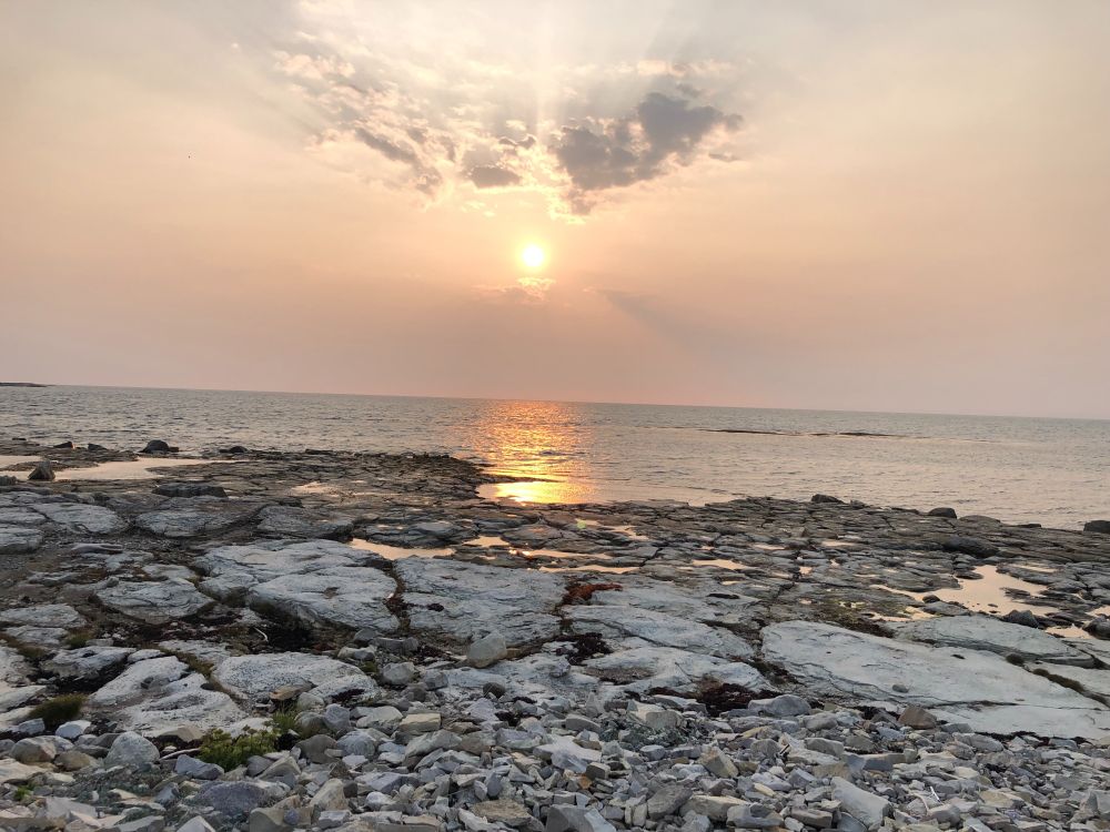 A gray flat-stoned shore, the sun approaching sundown in a pale orange-smoked sky, bright reflection on the gray sea.