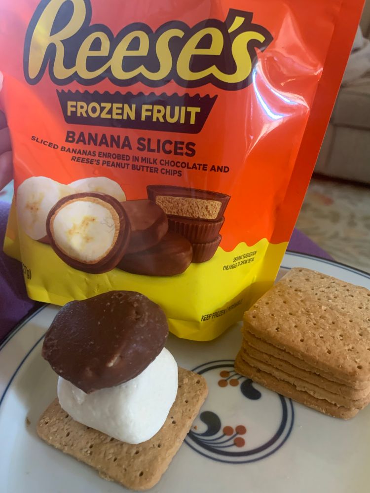Orange and yellow bag of Reese’s frozen fruit banana slices next to honeygram crackers stacked next to a frozen s’more comprised of a honeygram cracker, jumbo marshmallow and chocolate and peanut butter-covered banana slice, all on a plate.