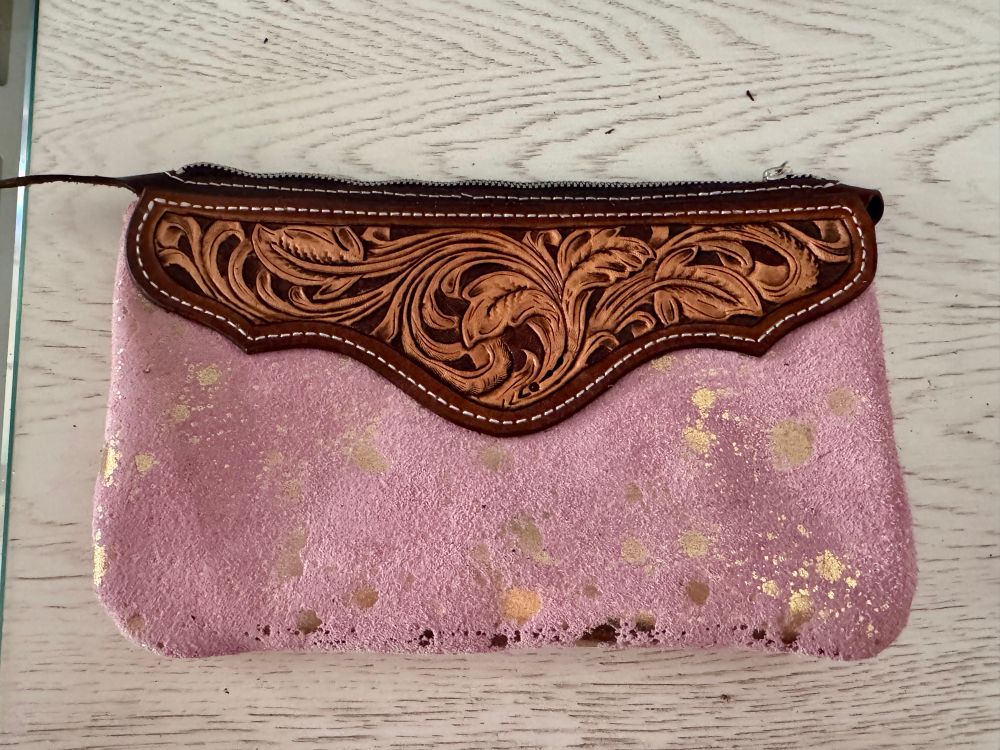 Image is of the back of a small handmade leather wristlet bag. It has a tooled scalloped panel and the body is baby pink suede that has a metallic gold ink print on it. The pouch zips at the top, and the tooling is floral in nature and features and spiraling vines and leaves. 