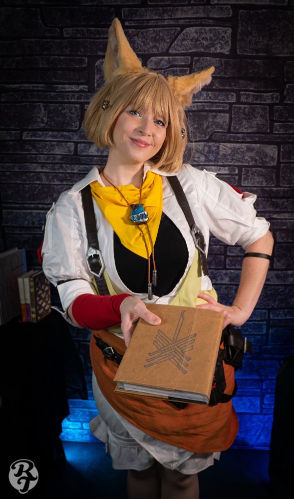 Image of a white woman in cosplay as Khloe Aliapoh from Final Fantasy 14.  She is wearing a white collared shirt and green overalls with leather straps. She has white bloomers with and orange sash and several leather pouches around her waist. She has a yellow bandana and a bolero tie like necklace with large turquoise gem. She has a short blonde bob wig and yellow cat ears. 

She is smiling as she holds out a book wrapped in brown paper towards the viewer. 