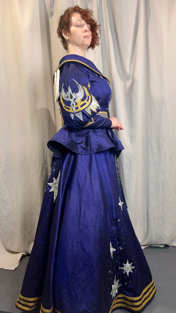 White woman with red hair posing in front of a grey back drop. She is wearing the FFXIv level 100 Astrologian gear. A jacket with bishop sleeves and oversized collar, and a line skirt. Both are made of blue silk fabric with silver embroidered stars and gold trim. Side view a large silver and gold embroidered circular design is bible on the right sleeve. 
