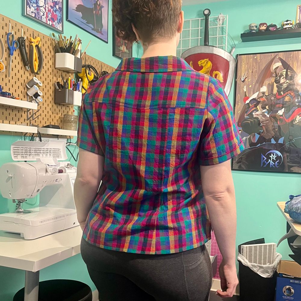 Back view image  of a white woman wearing a cropped button up t-shirt in a jewel toned multi colored  heck fabric. In the background there is sewing equipment. 