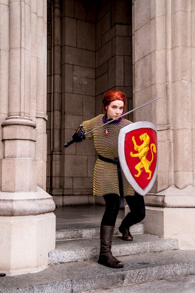 Cosplay photos. A red headed white woman wearing a gold chain mail shirt, black pants and brown clad high boots. She is wielding a sword and a shield painted with a red background and gold rearing lioness. The background is a stone building with gothic architecture. 