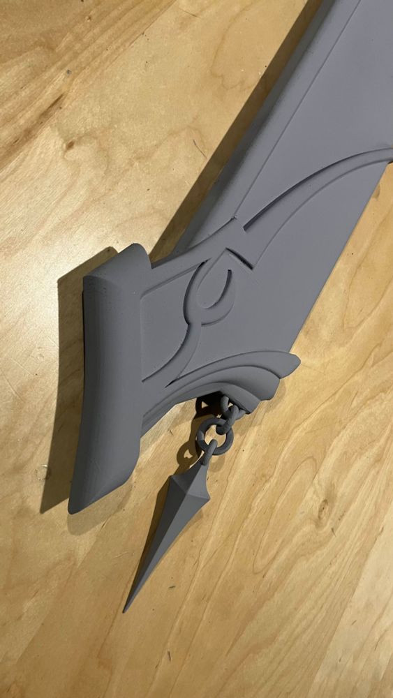 Photograph of a 3D print of the Astrologian card holder from FFXIV. Close up on the bottom section showing the bevels, and bottom dangling chain. The print is painted in grey primer and resting on a wooden table.