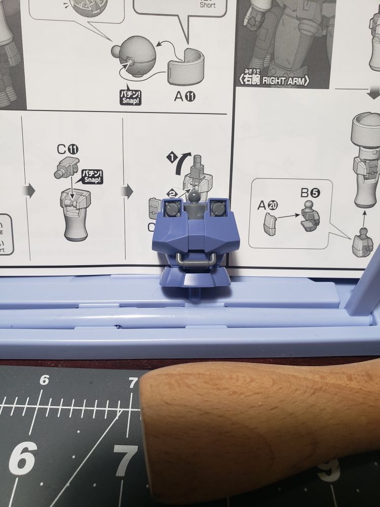 Plastic robot model torso on an instruction sheet.  Robot is light purple with gray clavicle.
Foreground is a cutting mat with a wooden handled brush.
