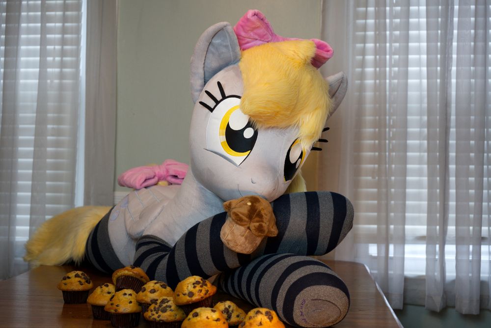 Lifesize Derpy plush wearing striped socks and pink bows on her head and tail. She is lying among piles of muffins and holds a plush muffin in her mouse magnetically.