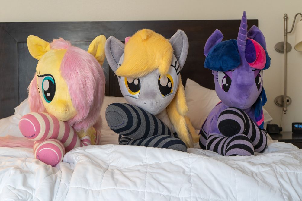 Three lifesize pony plushies, Fluttershy, Derpy, and Twilight. They lie comfortably on a bed with their front legs crossed.