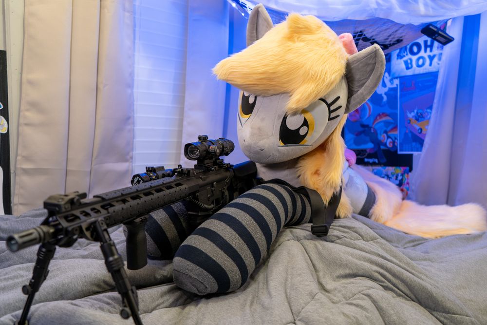 Lifesize Derpy plush wearing striped socks and pink bows on her head and tail. She lies on a bed and is staged to appear to be looking down the sights of a tactical-looking rifle.