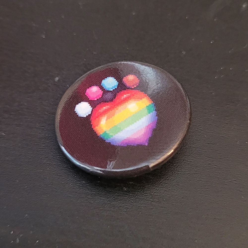 Pin badge with a print of a pixel art paw print in the shape of a heart, sporting the colors of the rainbow flag.