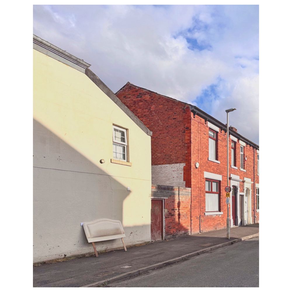 A street scene featuring a row of houses. The building on the left is cream-coloured with a single window visible. A white headboard leans against its wall on the pavement. The houses on the right are red brick terraces with a lamppost in front of them. A street sign with a speed limit of 20 is visible on the lamppost.