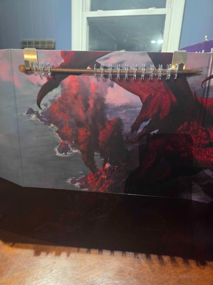 The front of an old Dungeons & Dragons dungeon master screen, with a pair of hanging hooks adhered to the front. The hooks are holding a wooden knitting needle which has plastic bobbins threaded along its length to create a sort of abacus-like setup.