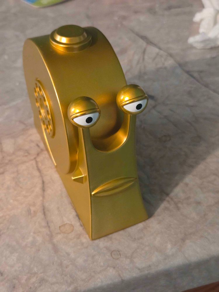 photo of a small plastic golden snail with a button on top. it's a "buster call" snail from the manga/anime One Piece.