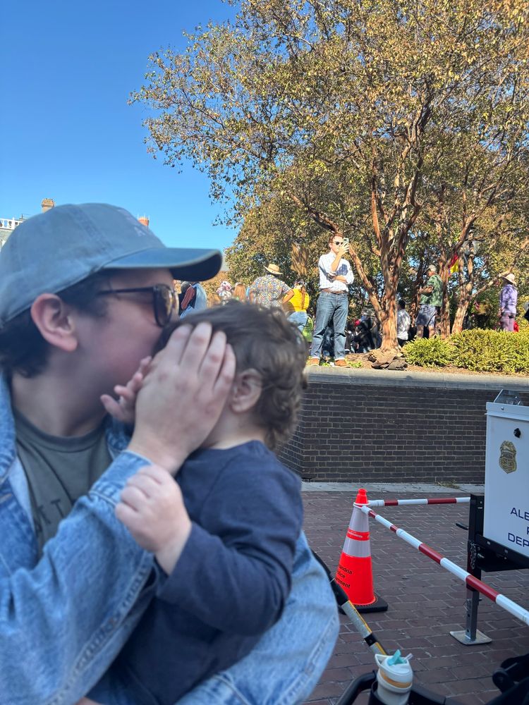 Man in baseball cap and denim jacket is outdoors holding a baby, covering the baby’s face. In the background, Andrew Egger is standing atop a brick wall on a video call, presumably the Bulwark’s livestream. 
