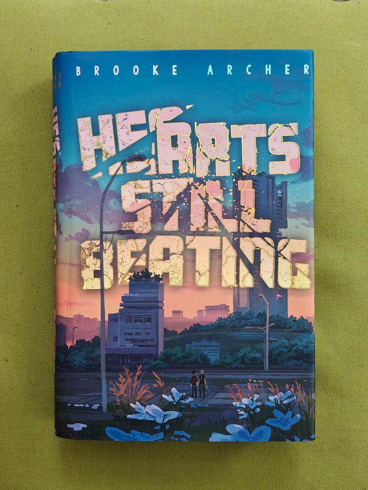 A cover of a post apocalyptic world and 2 girls holding hands.
Titled: Hearts Still Beating
By: Brooke Archer