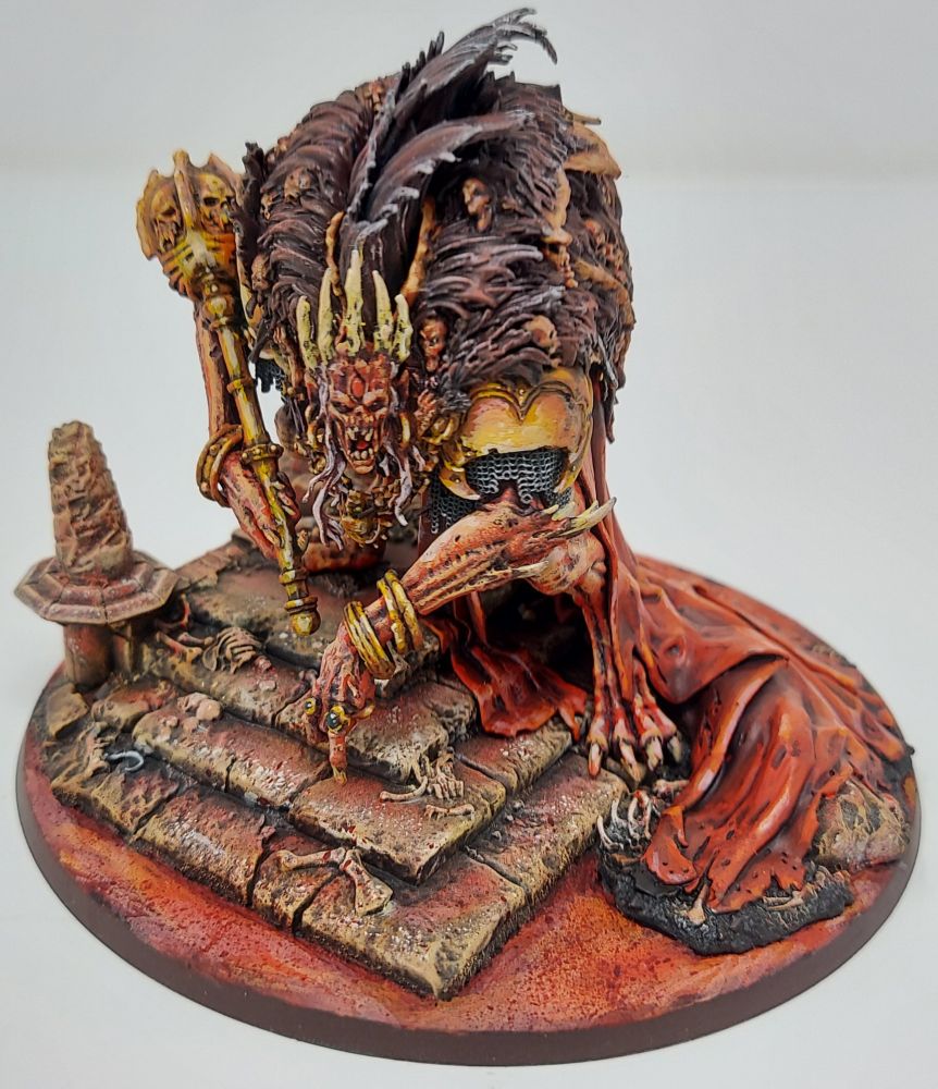 A miniature model of a monstrous, hunched, and ghoulish vampire king that is standing menacingly on top of a crumbling set of ruined stairs overlooking a lake of swirling blood. The ruin is littered with bones and debris, with the vampire's blood red cape and loincloth draped over the steps. Bright golden bangles hang from the creature's thin wrists as it holds a brilliant golden skeletal scepter in the crook of its long and corded arms. The creature stares at the viewer with beady, white eyes deep-set into a thin skeletal face, with a blood red gem embedded in its forehead and five bony spikes that protrude from its scalp, giving the impression of a morbid crown fused into its head. The creature wears several massive pelts over its hunched back, with gibbering skulls and screaming faces of the dead and insane tangled within the long and matted dark purplish brown fur. The model is extremely warm in tone, consisting majorly of off-white bone colors, blood reds, and vibrant orange and yellows for the golden adornments, with no blues or greens to be found. The coloration gives the impression of a cloying heat, an uncomfortable atmosphere of humidity born from the blood lake, with the smells of iron and rancid meat hanging ever present in the air.
