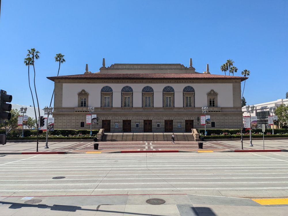 Pasadena Civic Auditorium. A multi-story building, done in Mediterranean revival style. There are few palm trees on each side.