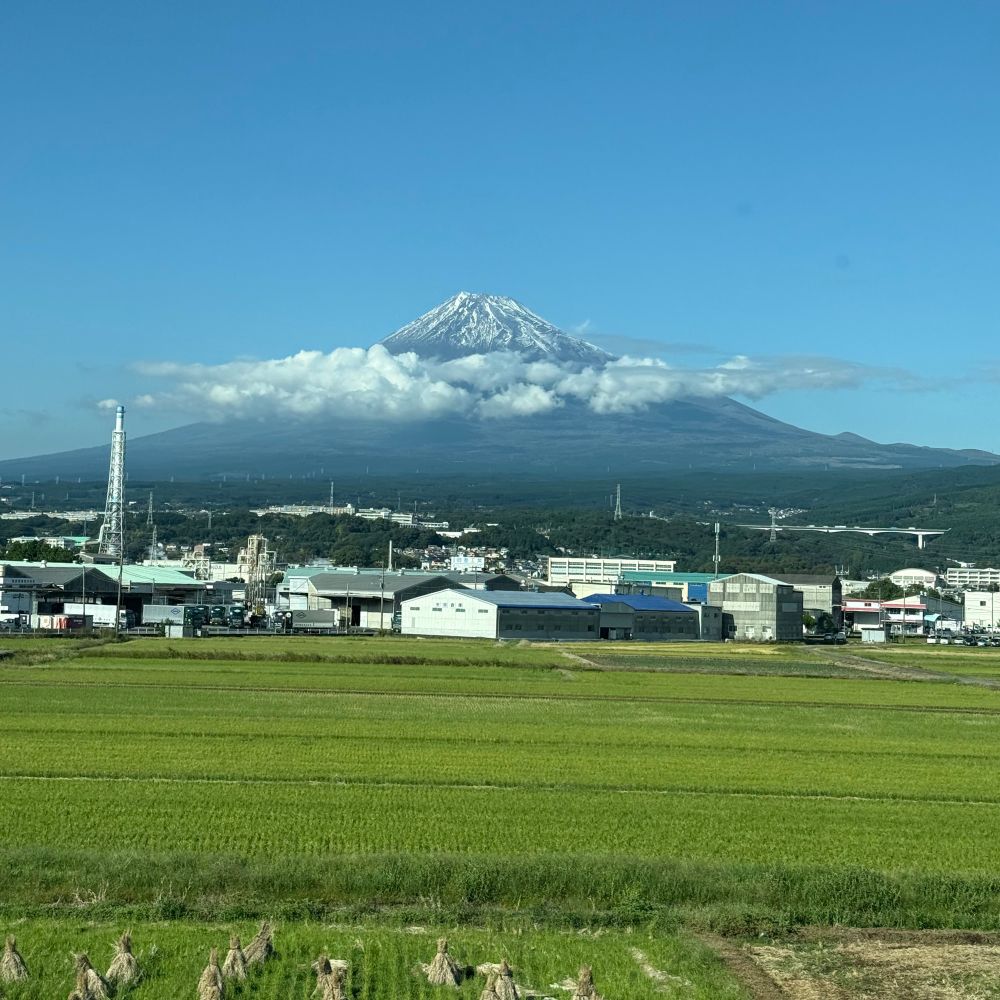 A photo of Mt. Fuji with light cloud coverage around the base of the mountain, with farmland in the foreground and a clear blue sky