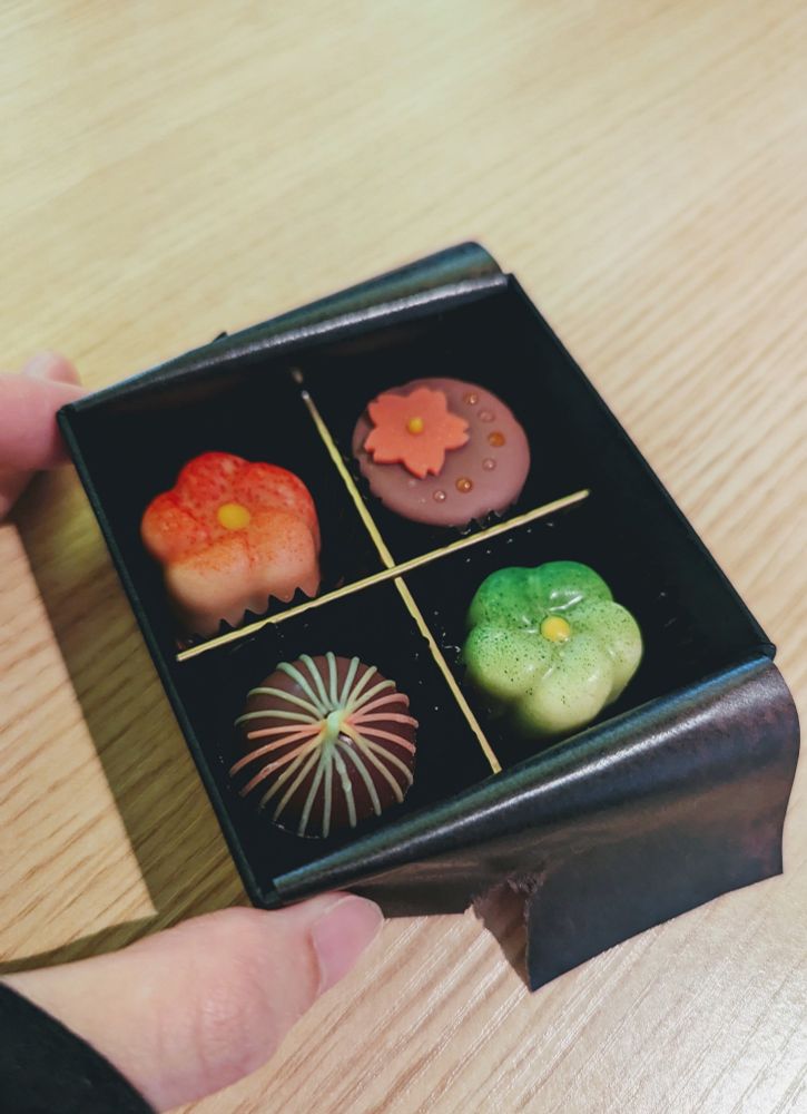 チョコレートの箱を手で持つ写真。
Chocolat Bel Amer 京都別邸の彩チョコレート4個入り。黒い小箱が金色の仕切りで4つに区切られており、手前から反時計回りに、手毬のようなチョコレート（抹茶キャラメル）、緑の梅の花のようなチョコレート（ピスタチオプラリネ）、円筒形の天面にピンクの花の形がのったチョコレート（白桃ガナッシュ）、赤の梅の花のようなチョコレート（シトロンプラリネ）。