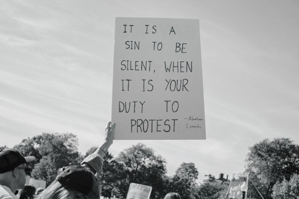 Another black and white photo from the same protest. This time from below eye level looking up at a sign held high. From this angle it appears as if the sign is above the tree line. On the sign is the Abraham Lincoln quote, “IT IS A SIN TO BE SILENT WHEN IT IS YOUR DUTY TO PROTEST” 