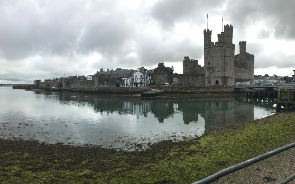 Tall turrets of a castle with a bridge on one side then the buildings of a town, but castle walls extend beyond.  All in front of a river or estuary.  An overcast sky.