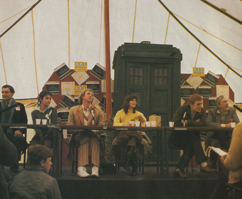 A convention panel from the 1980s.  From left to right: Anthony Ainley, Janet Fielding, Peter Davison (in costume), Sarah Sutton, Mark Strickson, and a bearded man I can't identify.  They are seated in what looks like a tent in front of boards with the BBC logo and a Tardis prop.