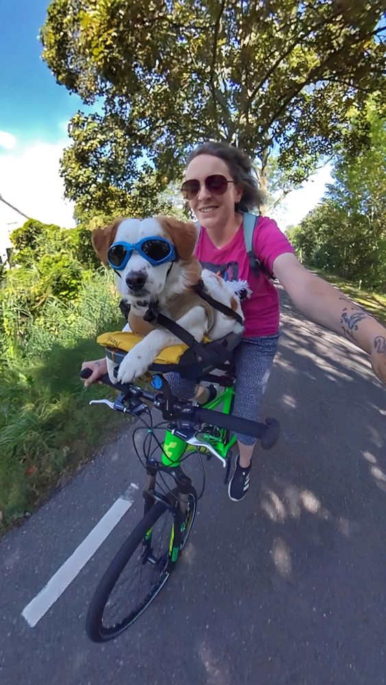 Woman with pink t-shirt riding in a green environment on a green bike with a little white dog with brown ears and spots chilling in a Buddyrider dog seat at the front 