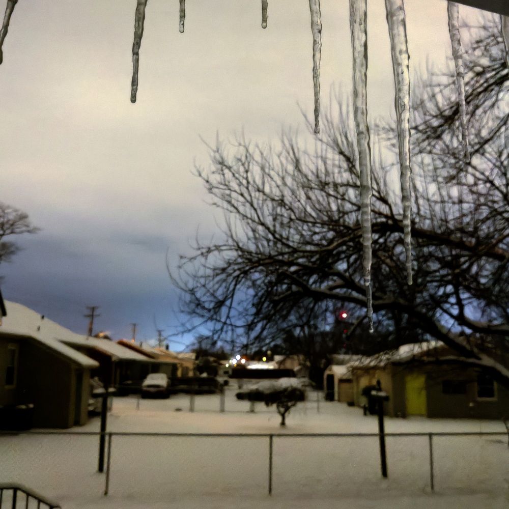 Night photo of icicles outside my bathroom window. They're in focus and the houses in the background are blurred. You can also see the snow in the backyards. ❄️