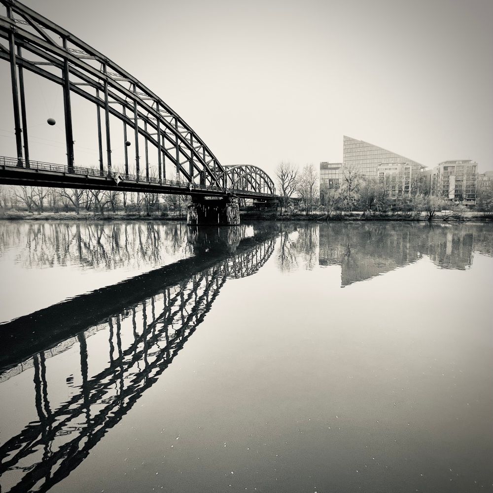Ein Schwarz-Weiß-Foto zeigt eine Stahlbrücke über einem ruhigen Fluss, in dem sich Brücke und Gebäude am anderen Ufer klar spiegeln. Die Szene wirkt ruhig, fast melancholisch.