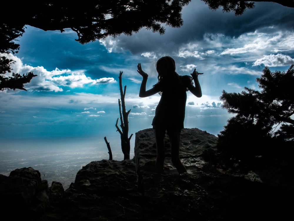 A young girl stands on a rock outcrop atop a mountain, overlooking a hazy city. Her figure is in silhouette and she is posed with hands in fun positions at shoulder height. She is back-lit by a blue sky with white clouds. 
