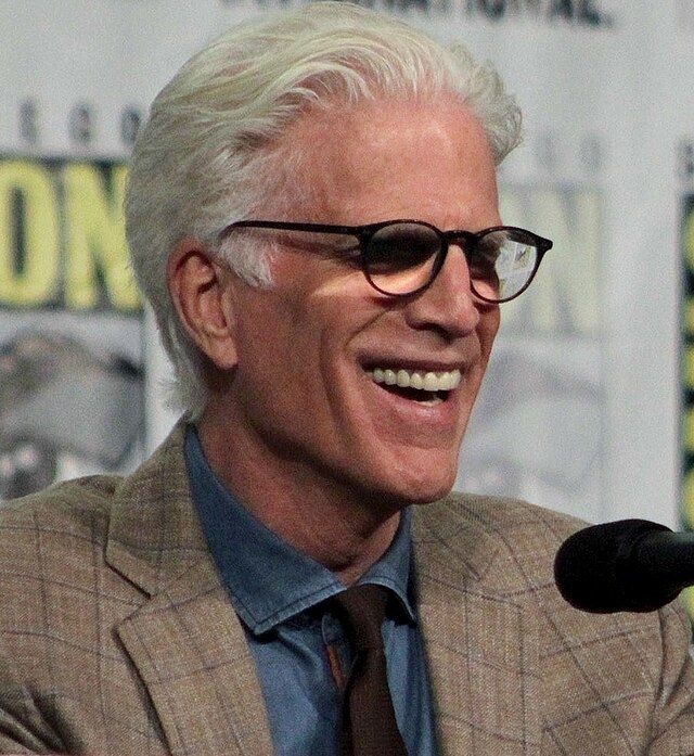 A picture of Ted Dason. He is an old man with neat white hair and glasses. He has a beige coat over a navy blue shirt and a maroon tie. He is standing behind a San Diego comicon panel and in front of him there is a mic.