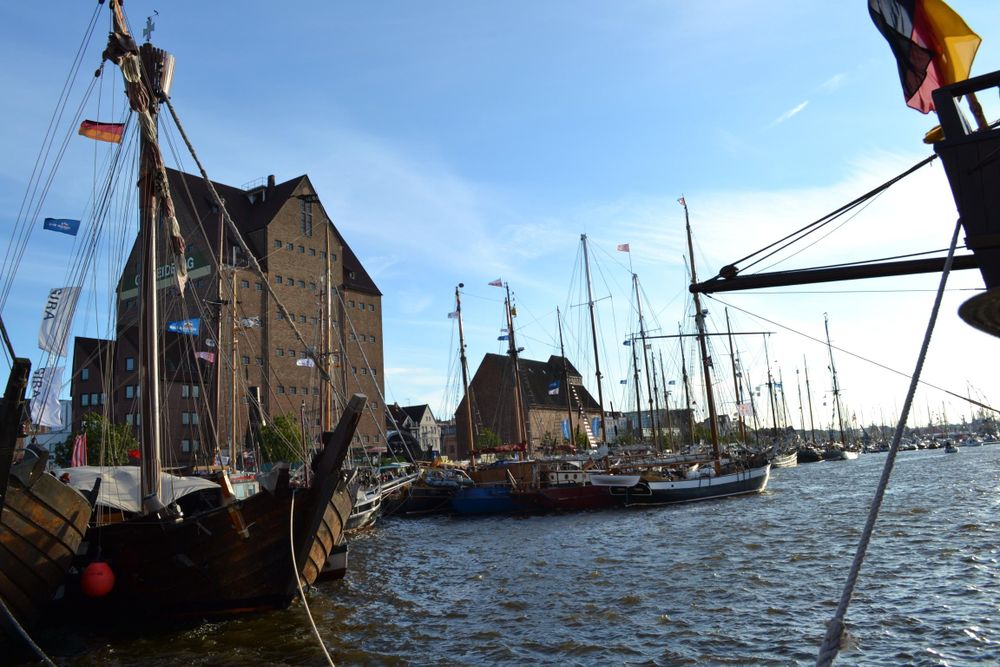 Hafen, im Hintergrund Lagerhäuser, im Vordergrund Hafenbecken mit Segelschiffen.