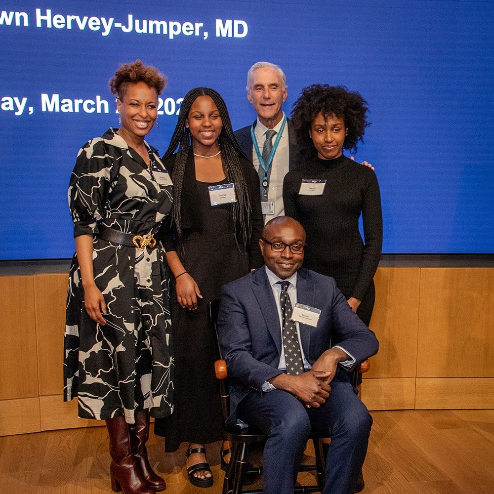Shawn Hervey-Jumper, MD, sitting in his chair with Heather Hervey-Jumper, MD (his wife), his daughters, and Mitchel Berger, MD, standing around him.