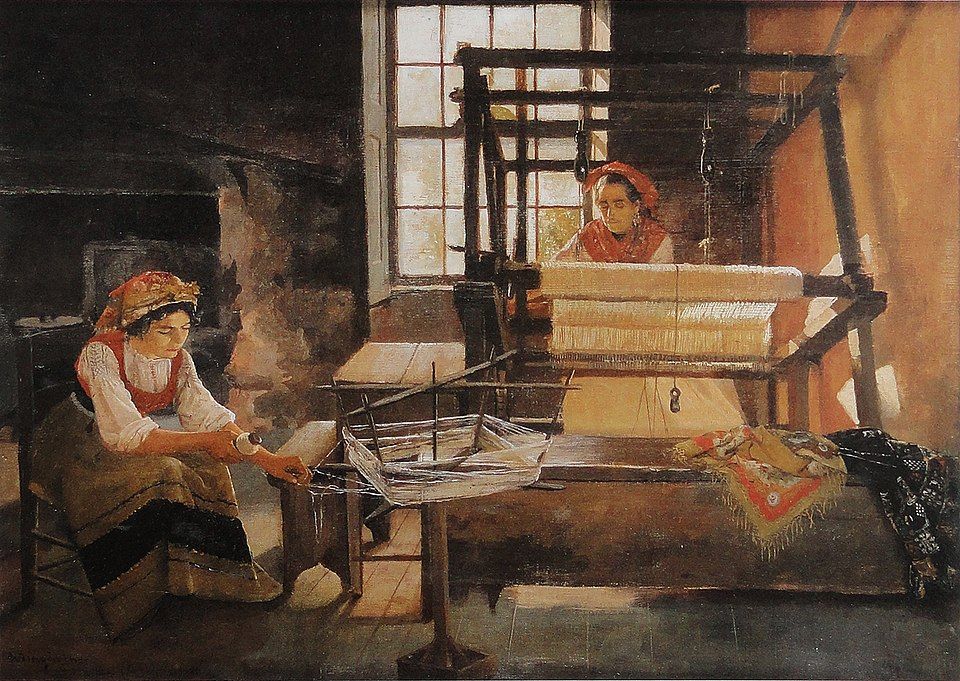 In the light of a large window in an otherwise dark room, a dark-haired woman is working on a large dark-framed loom. She is looking down at the fabric she is weaving and is obviously intent on her work. Her hair is covered by a red scarf and she has another red scarf arranged over her white top and is wearing a white skirt or white apron. A younger woman, also wearing a red scarf over her dark hair, is seated to the side of the loom winding thread. She is dressed in a red bodice over a white blouse and a light brown skirt with a wide black stripe above the light brown ruffle. There is a bench in front of the loom upon which a fringed red shawl and a black shawl have been thrown. Image by Pedro Weingärtner (1853–1929) from Wikimedia Commons.