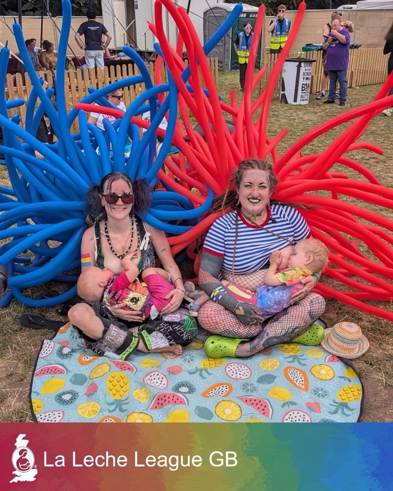 Two mums breastfeeding while sitting cross-legged on a colourful picnic blanket at a festival on a warm day. They each have a large bunch of long balloons tentacles tied to their backs, one in red, the other in blue. No text